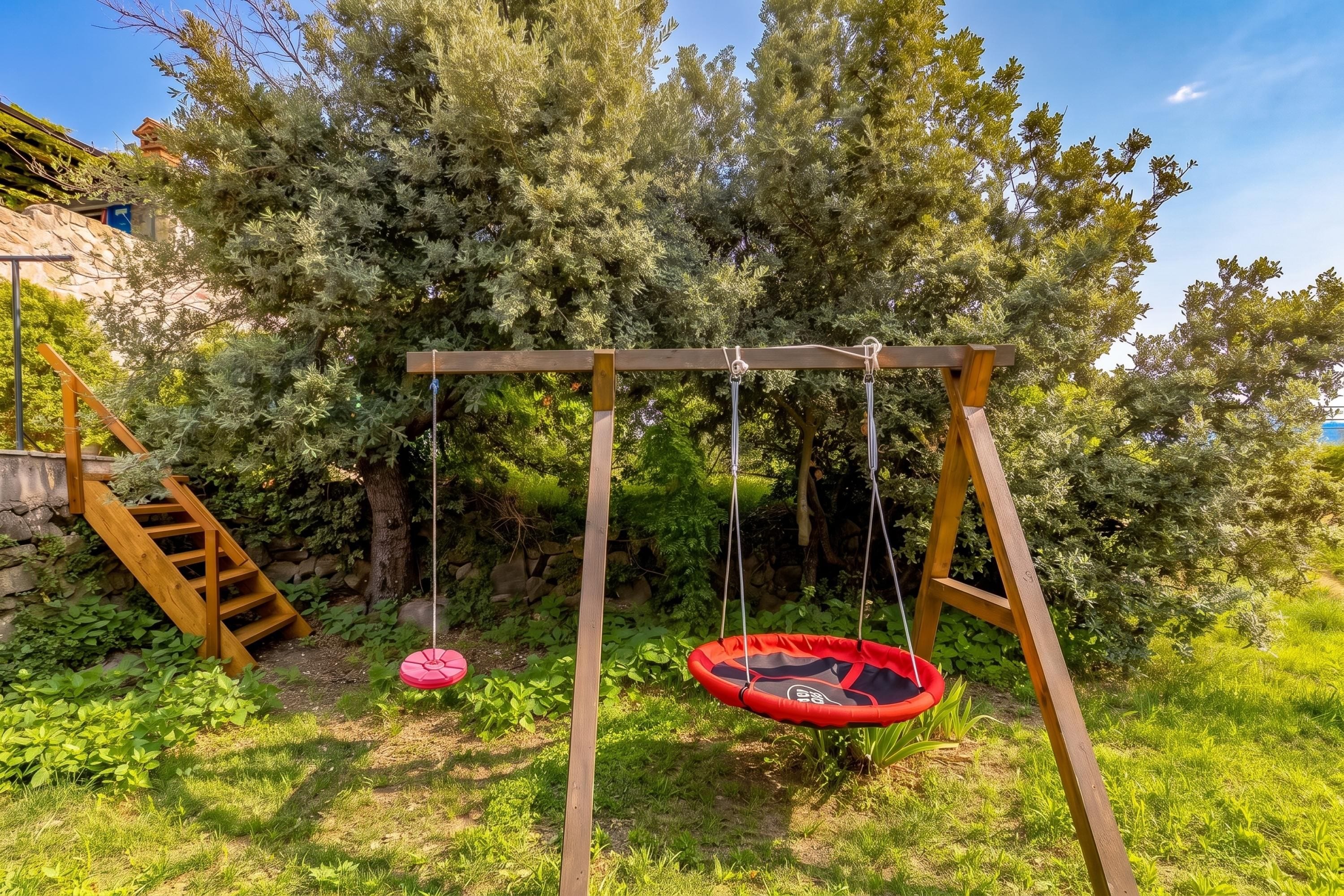Un columpio de madera con dos asientos, uno rojo y uno rosa, en un jardín con un muro de piedra y árboles.