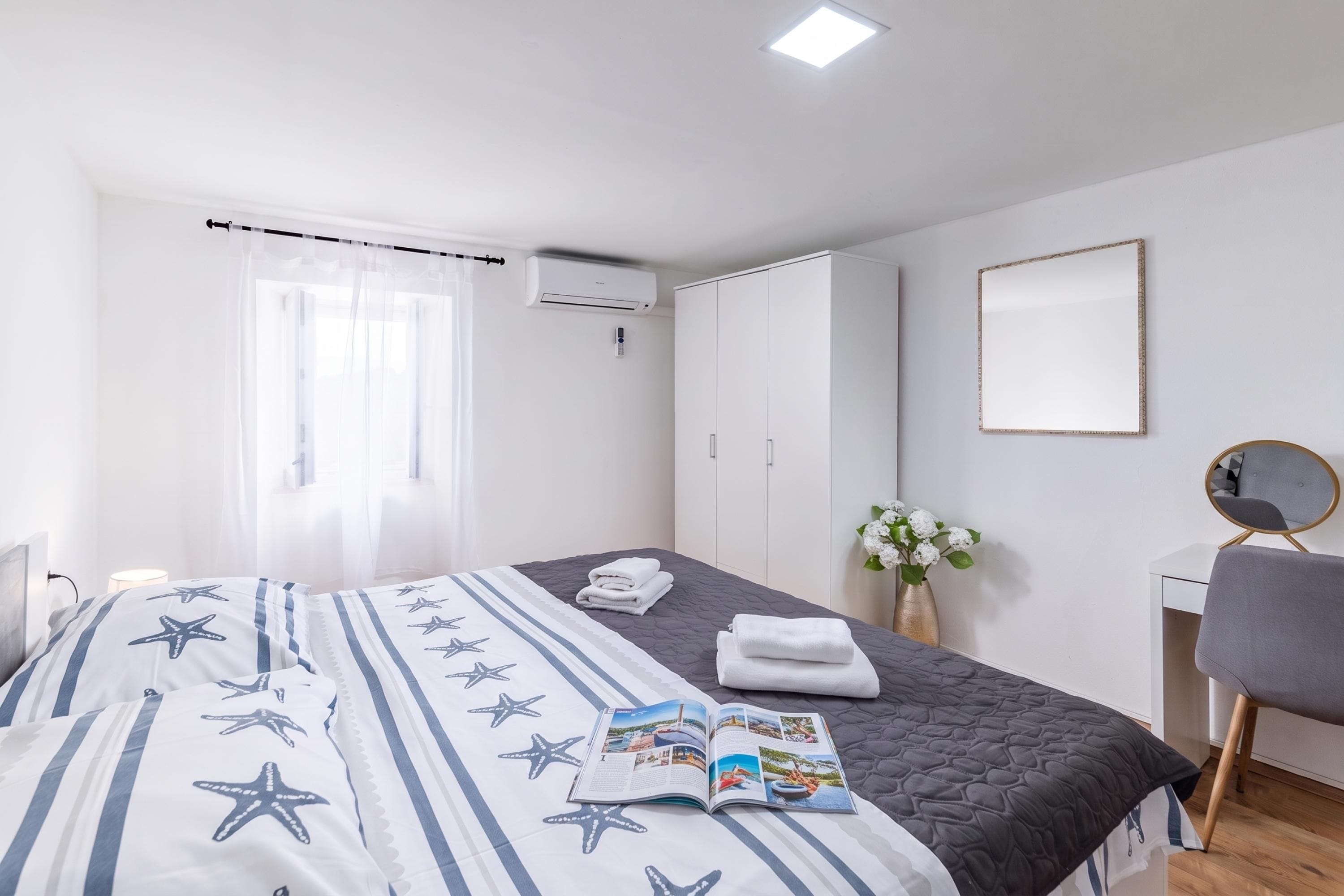 A hotel room with a bed, a bedside table, a chair, a desk, a mirror, a window with curtains, an air conditioning unit, and a wall-mounted mirror.