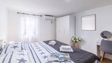 A hotel room with a bed, a bedside table, a chair, a desk, a mirror, a window with curtains, an air conditioning unit, and a wall-mounted mirror.
