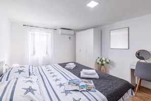 A hotel room with a bed, a bedside table, a chair, a desk, a mirror, a window with curtains, an air conditioning unit, and a wall-mounted mirror. - Hidden Hill Lopar With Sauna & Jacuzzi (Lopar)