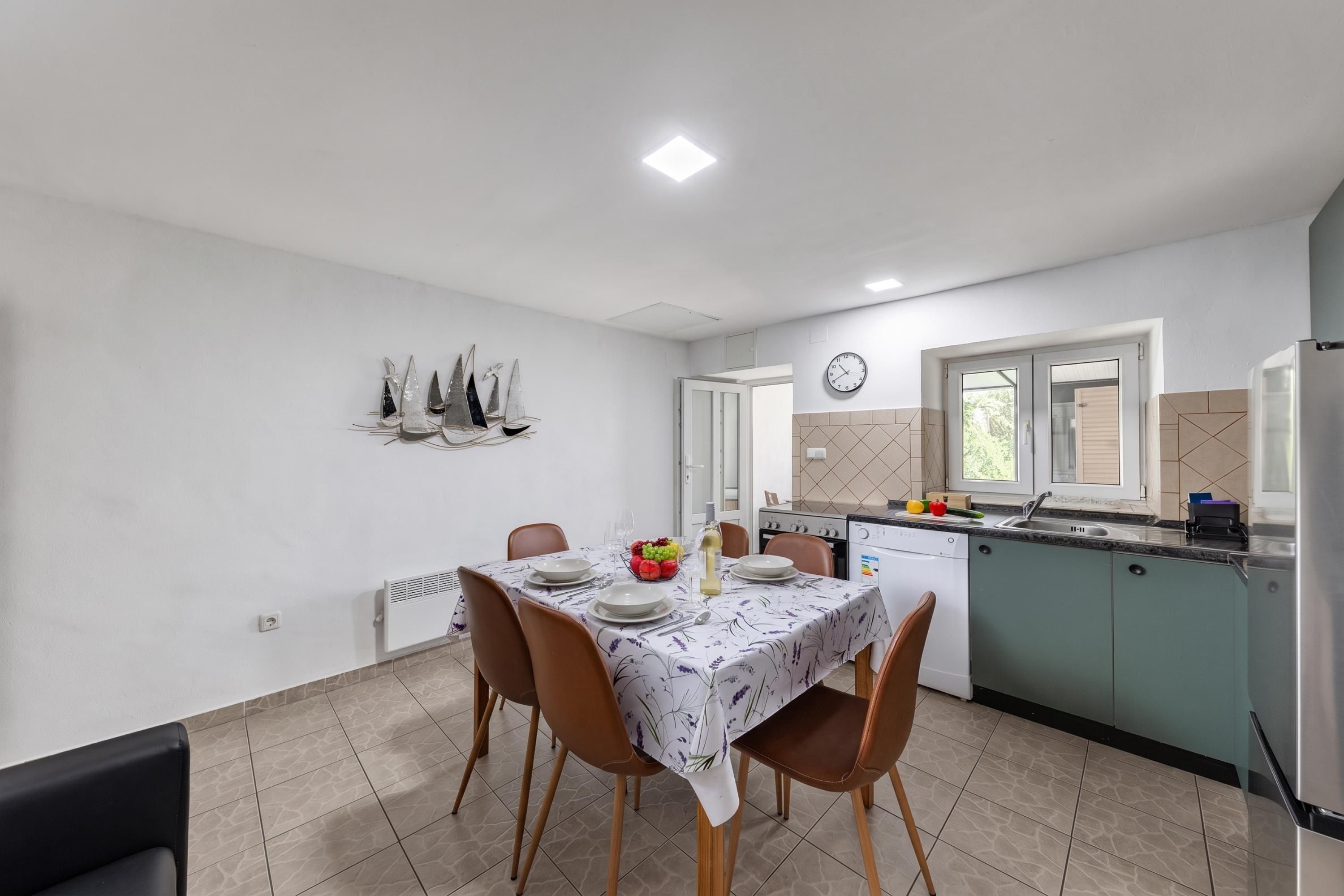 Un comedor con mesa preparada para cuatro, una cocina con electrodomésticos modernos y un reloj en la pared.