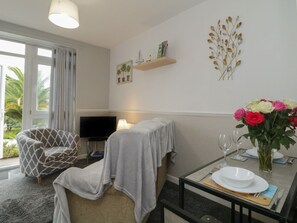 A hotel room with a sofa, a dining table with plates and glasses, a TV, a window with curtains, and a wall with a plant and decorative elements. - Flat 2, Big Tree Holiday Flats, Paignton (Paignton)