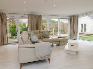 A modern living room with a beige sectional sofa, a white coffee table, and large windows offering a view of greenery outside. - Priory House, Barnstaple (Barnstaple)