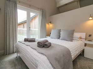 A bedroom with a large bed, a nightstand, a lamp, and a view of a wooden deck outside. - Priory House, Barnstaple (Barnstaple)