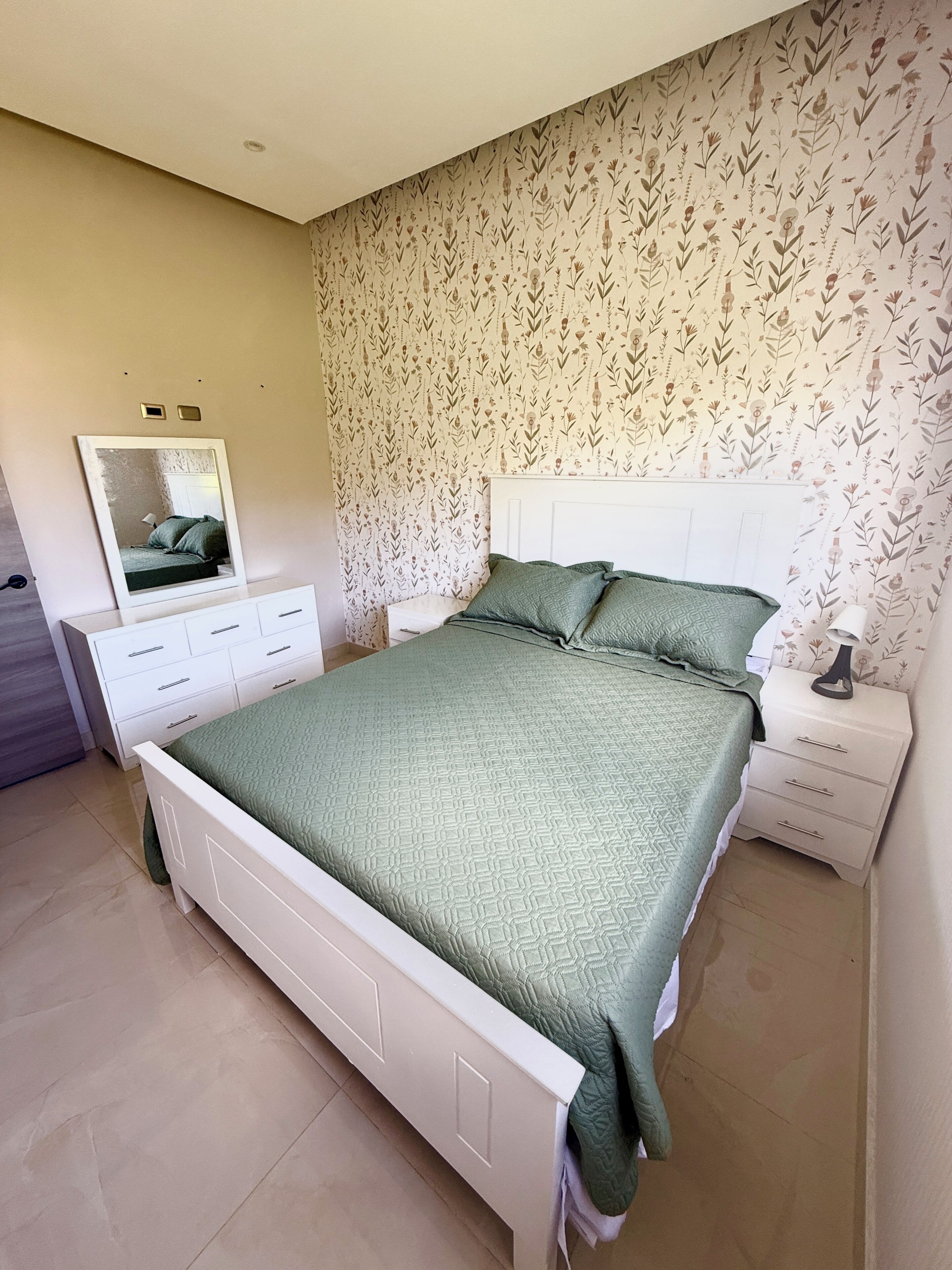 Quarto com cama, cômoda, mesa de cabeceira, espelho e papel de parede floral.