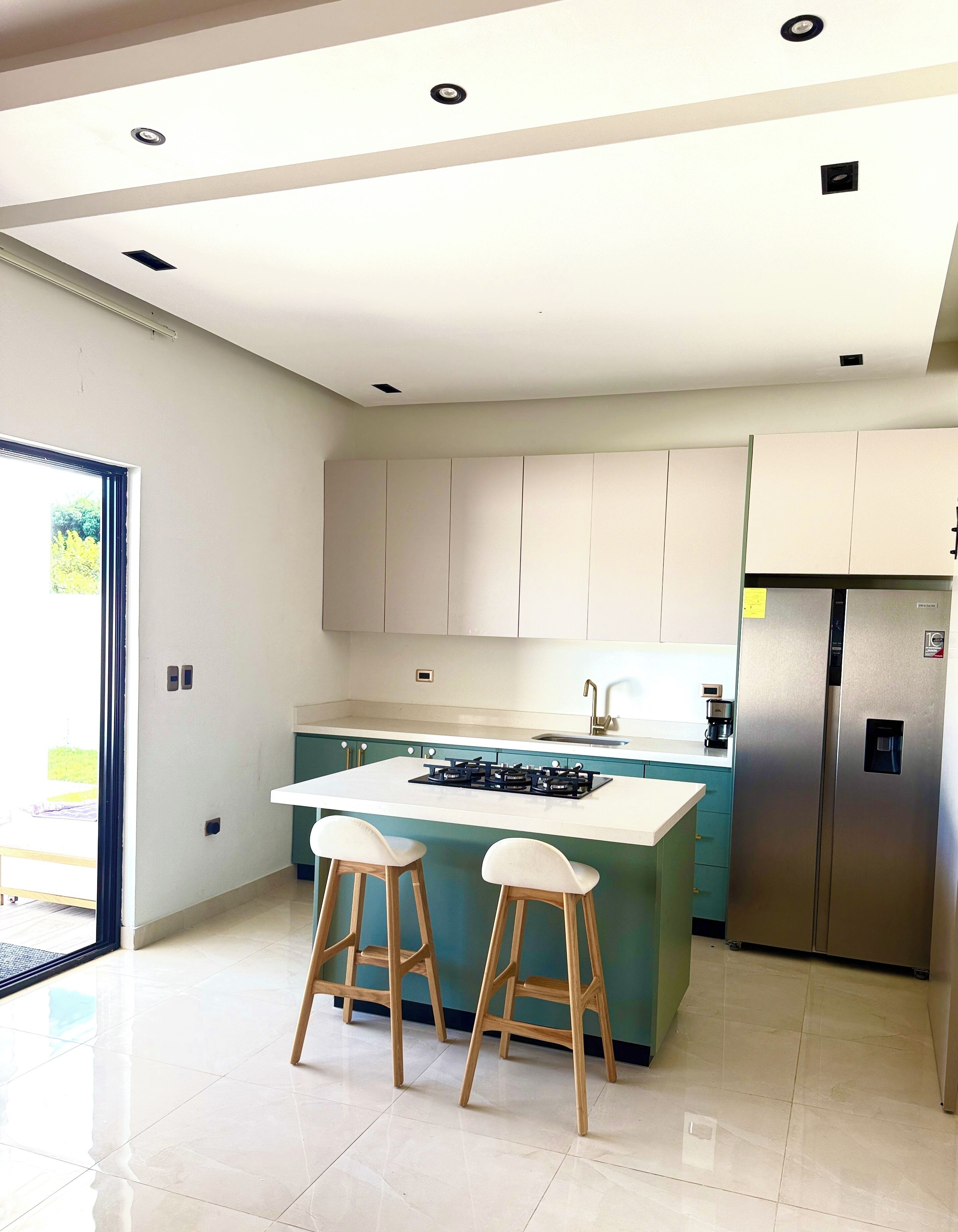 A modern kitchen with a central island, bar stools, a stainless steel refrigerator, and recessed ceiling lighting.