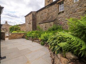 A stone building with a paved courtyard, surrounded by lush greenery and ferns. - Rose Tree Cottage, Skipton (Skipton)