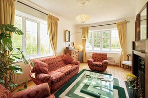 A living room with a sofa, armchair, and coffee table, large windows with curtains, a radiator, and a lamp.