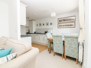 A living room with a sofa, a dining table with chairs, a kitchen in the background, and framed pictures on the wall. - Strawberry Cottage, Penryn (Penryn)