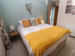 A bedroom with a bed, a bedside table with a lamp, a window with curtains, and a wall-mounted decoration. - Blake Lodge, Crantock (Newquay)