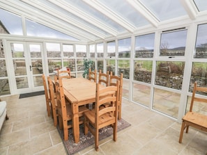 A conservatory with a wooden table and chairs, large windows, and a view of the countryside. - Hillside Farm, Kirkby Stephen (Kirkby Stephen)