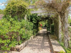 Ein Gartenpfad mit einer hölzernen Pergola, umgeben von üppigem Grün und blühenden Pflanzen.