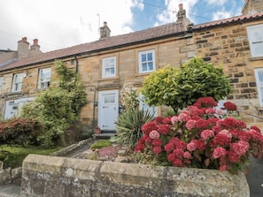 A stone building with a red-tiled roof, a white door, and a garden with red flowers and shrubs. - Manor Cottage, Cloughton (Scarborough)