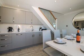 A modern kitchen with grey cabinets, a white countertop, and a dining area with a table set for two. - Charming Sandunes 2 Retreat in Camber (Rye)