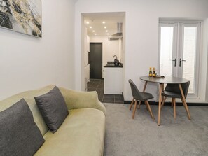 A living room with a sofa, a dining table with chairs, a kitchen area, and a door leading to another room. - Flat 1, Mona House, Deganwy (Conwy)