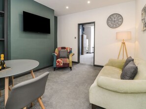 A living room with a TV, a sofa, a chair, a lamp, and a clock on the wall. - Flat 1, Mona House, Deganwy (Conwy)
