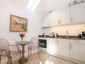 A modern kitchen with white cabinets, a round table with chairs, a painting on the wall, and a vase with red flowers. - The Garden Room in Chichester. (Chichester)