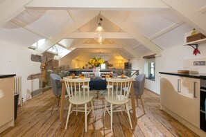 A dining area with a table set for two, wooden chairs, a hanging light fixture, and a kitchen in the background. - Stable Loft, Watergate Bay (Newquay)
