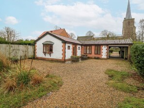 A building with a white facade and red brick accents, a gravel driveway, and a church tower in the background.