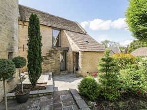 A stone house with a sloped roof, a small garden with potted plants, and a clear sky. - The Barn, Colerne, Wiltshire (Chippenham)