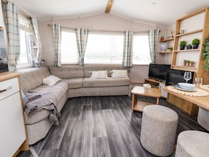 A cozy living area with a sofa, a coffee table, and a TV. There are windows with curtains, a shelf with plants, and a wooden table with a glass. - Lawi39: Charming Retreat in Towyn (Abergele)