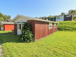 A red wooden building with a door and windows, surrounded by green grass and a hill.