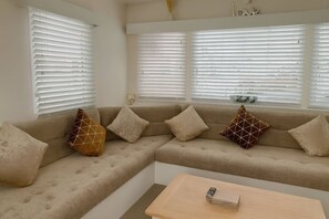 A living room with a beige sofa, multiple cushions, a small table, and large windows with blinds. - Golden Gate Holiday Centre - 19058, Towyn (Abergele)