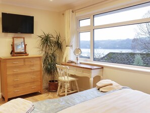 A bedroom with a bed, a desk, a chair, a dresser, a television, a window with a view of a lake, and a potted plant. - Treetops: A Hidden Gem in Golant (Fowey)