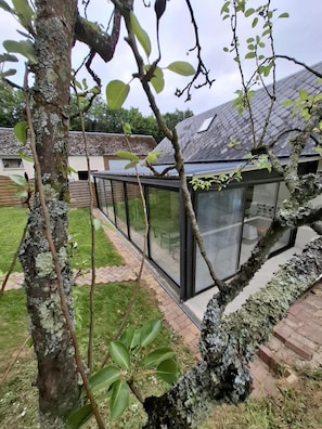 A modern building with large glass windows, a brick pathway, and a tree in the foreground. - Gîte Ressources: Jacuzzi, Private Terrace & Garden (La Frénaye)