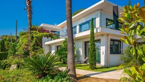 Una casa moderna de dos pisos con balcón, rodeada de abundante vegetación y palmeras.