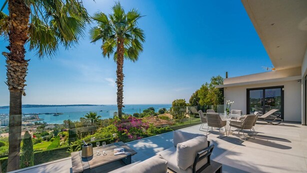 Un patio exterior moderno con vistas al mar, amueblado con sofás y una mesa, palmeras y una barandilla de cristal.