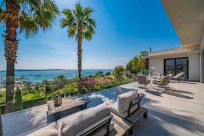 A modern outdoor patio with a view of the sea, furnished with sofas and a table, palm trees, and a glass railing. - Contemporary villa with ocean views and a pool, near Cannes (Vallauris)