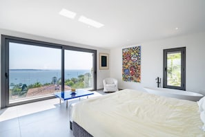 A modern bedroom with a large bed, a glass table, a white chair, a bathtub, and a view of the sea. - Contemporary sea view villa with pool, near Cannes (Vallauris)