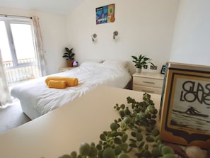 A hotel room with a bed, a bedside table with a plant, a window with curtains, and a wall with a poster. - Lowena Lodge, Millbrook (Torpoint)