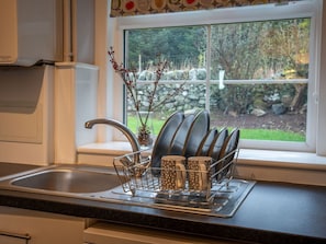 A kitchen with a sink, a dish rack with plates and mugs, and a window with a view of greenery and a stone wall. - The Bothy, Blairgowrie (Blairgowrie)