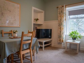 A room with a dining table, chairs, a television on a stand, a map on the wall, and a window with a curtain. - The Bothy, Blairgowrie (Blairgowrie)