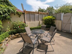 A patio with a round glass table and four chairs, surrounded by a garden with various plants and a wooden fence. - Scalby Grove Cottage, Scalby (Scarborough)