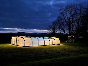 A large, illuminated greenhouse structure in a grassy field at night.