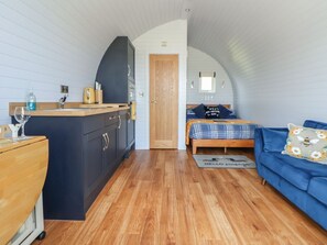A compact room with a kitchenette, a bed with a blue and plaid bedspread, a blue sofa with a floral pillow, and a wooden door. - Honeypot Hideaways - Manuka Retreat (Chester)