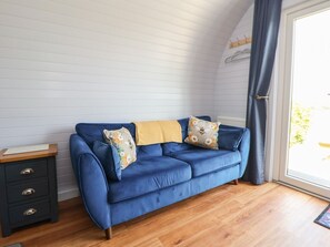 A blue sofa with floral pillows, a wooden side table, and a view through a large window. - Honeypot Hideaways - Manuka Retreat (Chester)