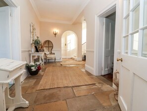 Un couloir avec une porte blanche, une table blanche, un miroir, un vase de fleurs, un tapis et une statue de paon.