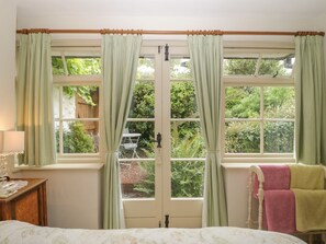A bedroom with a bed, a window with curtains, a bedside table with a lamp, and a view of the outdoors.