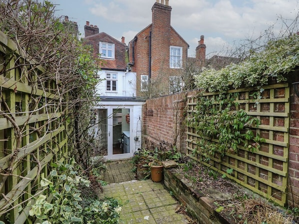 A narrow courtyard with a brick wall, a wooden fence, and a small door leading to a room.
