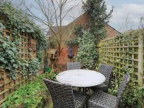 A round wooden table with wicker chairs in a garden setting with brick walls and climbing plants.