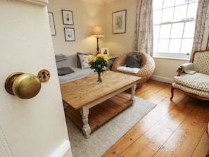A room with a wooden floor, a sofa, a wicker chair, a wooden table, and a door with a brass handle.