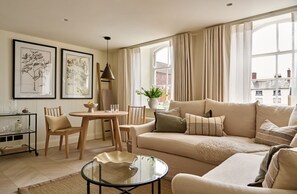 Ein Wohnzimmer mit einer Couch, Sesseln, einem Glastisch, einem Esstisch mit Stühlen, gerahmten Pflanzenbildern an der Wand und einem Fenster mit Vorhängen.