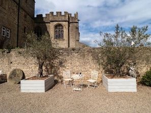 A courtyard with a stone building featuring a large arched window and a castle-like tower. There are two white planters with trees and a small table with chairs. - Catherine Parrs Sewing Room, Bedale (Bedale)