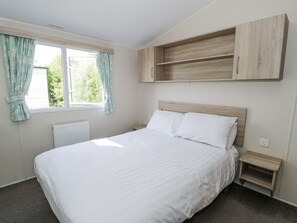 A bedroom with a bed, a window with curtains, a radiator, a wooden shelf, and a bedside table. - Manor House Gold 2 Bedroom (20C), Aspatria (Maryport)