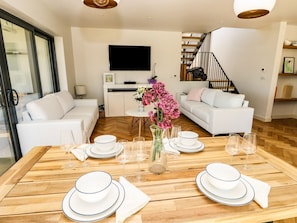 Un comedor con una mesa de madera dispuesta para cuatro, sofás blancos, un televisor y una escalera.