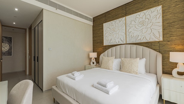 A hotel room with a large bed, two bedside tables with lamps, a chair, and a decorative wall panel. - Live the Vibe Upgraded 2BR JBR Coastal Living (Dubai)